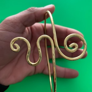 Può includere: Un bracciale in metallo color oro con un design decorativo a spirale. Il bracciale presenta una barra verticale e due accenti curvi a spirale. Il pezzo è tenuto su uno sfondo verde.