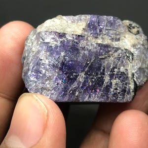 May include: A close-up of a rough-cut, natural gemstone. The stone is a deep purple color with areas of white and gray. The surface has a crystalline texture with iridescent flashes of color.