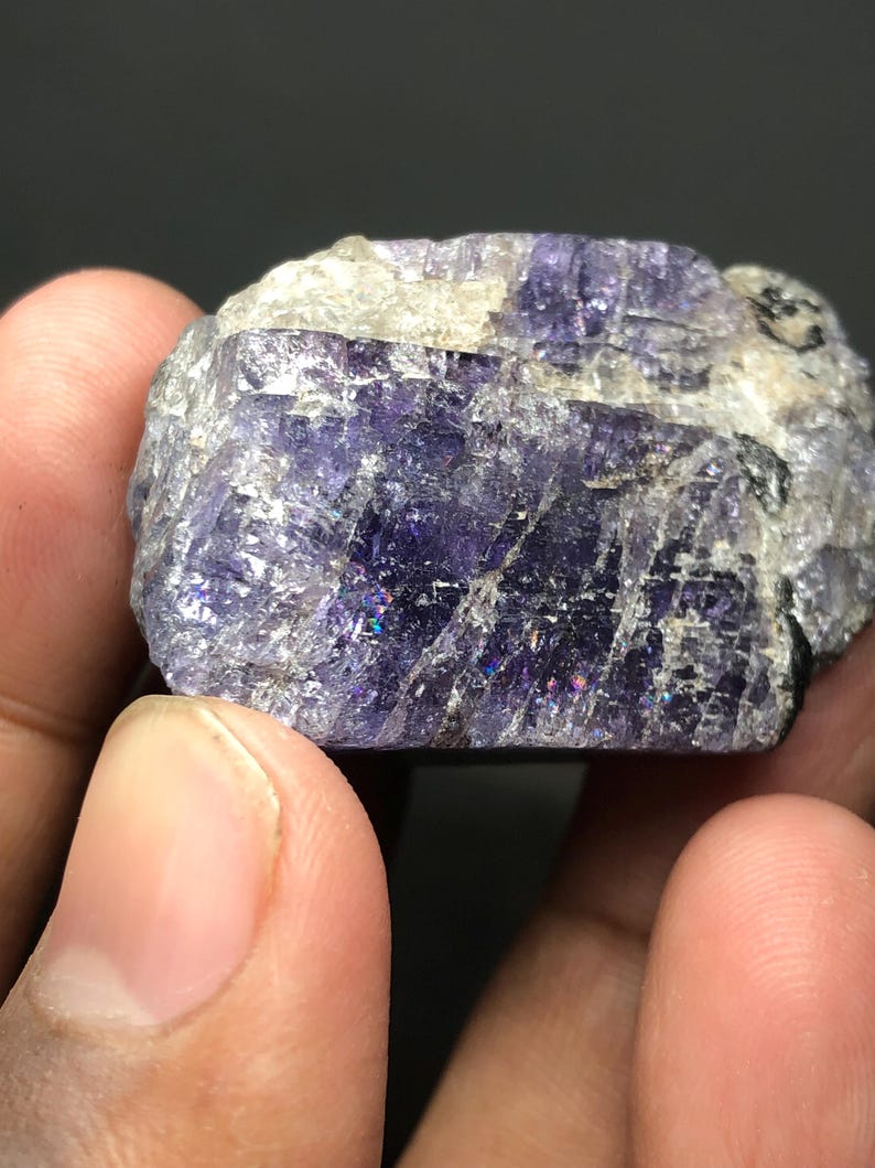 May include: A close-up of a rough-cut, natural gemstone. The stone is a deep purple color with areas of white and gray. The surface has a crystalline texture with iridescent flashes of color.