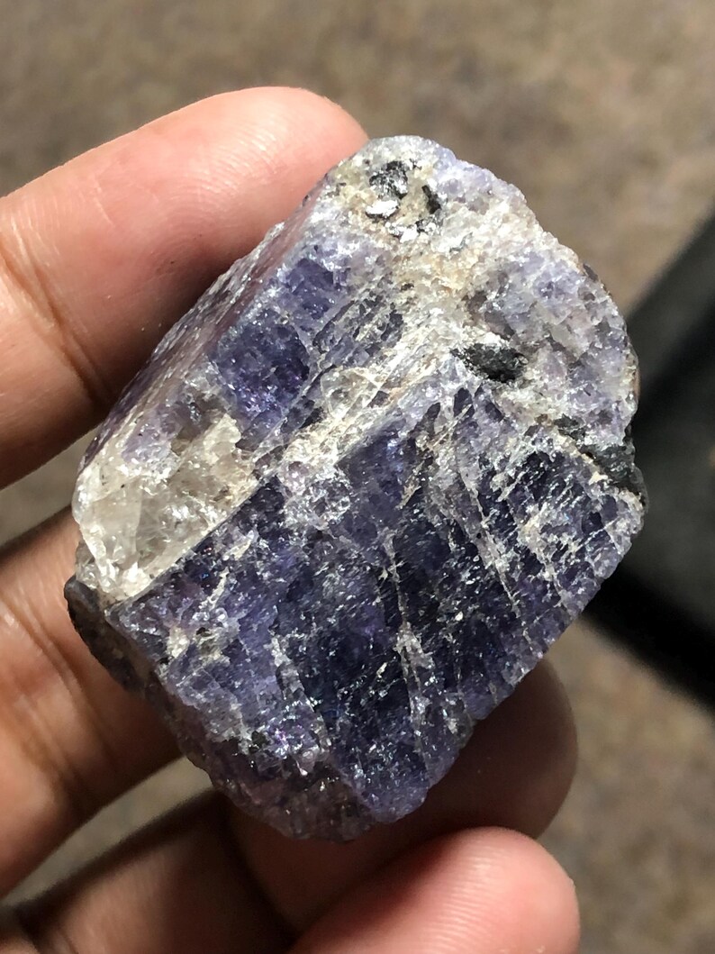 May include: A raw, uncut gemstone with a deep indigo and lavender color. The mineral has a rough, textured surface with areas of white and black inclusions. The stone is held in a hand, showcasing its natural, unpolished state.