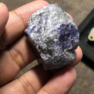 May include: A rough-cut, natural gemstone with a mix of gray and purple hues. The stone has a textured surface and irregular shape, showcasing its raw, unpolished state. The color ranges from deep violet to lighter shades, with areas of gray and silver.