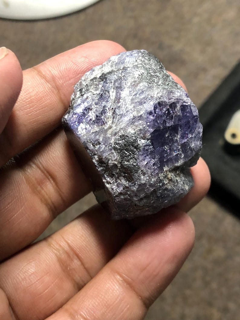 May include: A rough-cut, natural gemstone with a mix of gray and purple hues. The stone has a textured surface and irregular shape, showcasing its raw, unpolished state. The color ranges from deep violet to lighter shades, with areas of gray and silver.