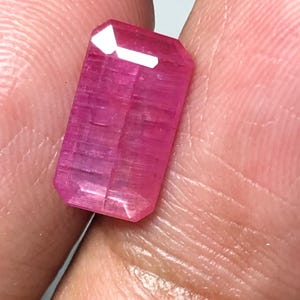 May include: A faceted, rectangular pink gemstone with a polished surface. The gem has a vibrant, saturated color and appears to be held between fingers. The gem's facets reflect light, creating bright highlights.