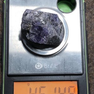 May include: A rough, dark purple gemstone rests on a digital scale. The scale displays a weight of 45.149 grams. The scale is black and silver, with the brand name "Brifit" visible.