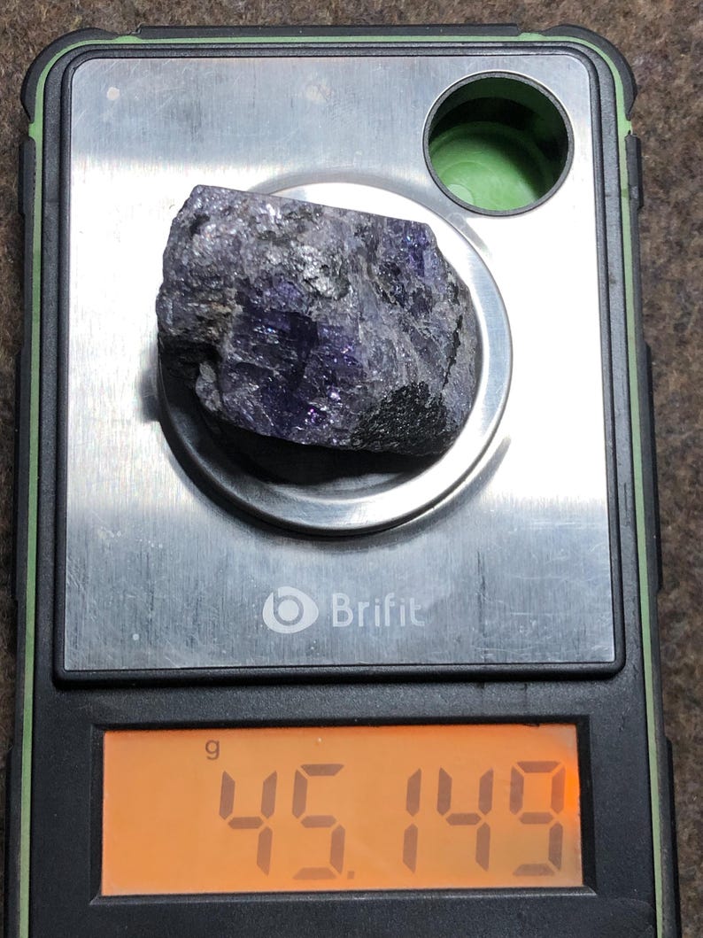 May include: A rough, dark purple gemstone rests on a digital scale. The scale displays a weight of 45.149 grams. The scale is black and silver, with the brand name "Brifit" visible.