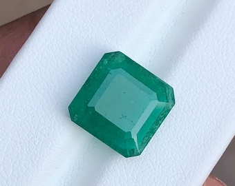 May include: A vivid, emerald-cut green gemstone. The gem is a deep, rich green colour with a rectangular shape and beveled edges. The gemstone is displayed on a white surface.