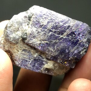 May include: A raw, natural mineral specimen with a mix of purple and gray hues. The crystalline structure displays a shimmering, iridescent effect. The rough stone is held in a hand, showcasing its texture and color variations.