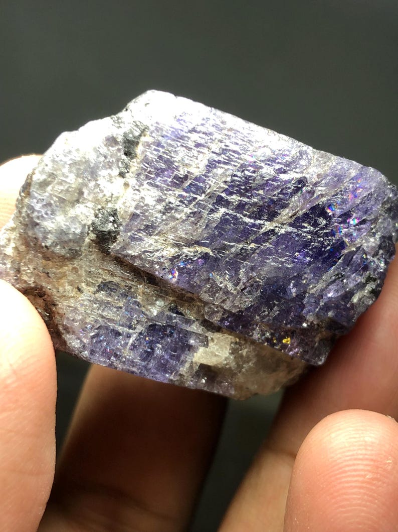 May include: A raw, natural mineral specimen with a mix of purple and gray hues. The crystalline structure displays a shimmering, iridescent effect. The rough stone is held in a hand, showcasing its texture and color variations.