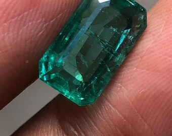 May include: A rectangular emerald-cut gemstone with a deep green colour and visible internal inclusions. The stone features a faceted surface that reflects light, showcasing a transparent quality and a polished finish with sharp, defined edges.