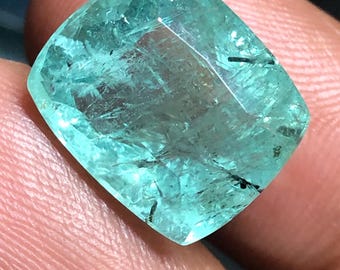 May include: A cushion-cut gemstone with a translucent light green colour and visible internal inclusions. The stone features a faceted surface that reflects light, showcasing a mix of clear areas and dark needle-like mineral deposits throughout.