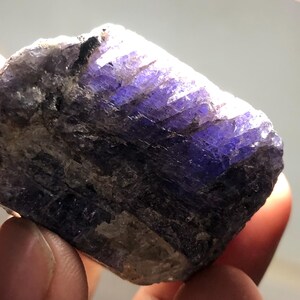 May include: A rough-cut, natural tanzanite gemstone. The stone displays a deep, vibrant purple hue with areas of blue and brown. The crystalline structure is visible, and the stone is held between fingers.