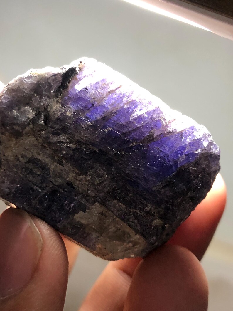 May include: A rough-cut, natural tanzanite gemstone. The stone displays a deep, vibrant purple hue with areas of blue and brown. The crystalline structure is visible, and the stone is held between fingers.