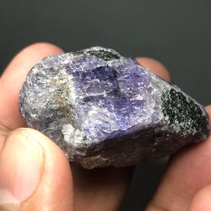 May include: A rough-cut, natural tanzanite gemstone. The stone displays a mix of colors, including deep purples, blues, and grays, with areas of black and a crystalline structure. The gemstone is held in a hand.