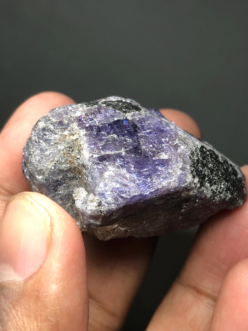 May include: A rough-cut, natural tanzanite gemstone. The stone displays a mix of colors, including deep purples, blues, and grays, with areas of black and a crystalline structure. The gemstone is held in a hand.