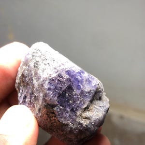 May include: A rough-cut, natural gemstone with a mix of gray and purple hues. The stone has a crystalline structure with visible facets and a textured surface. The color ranges from deep violet to lighter shades, with areas of gray.