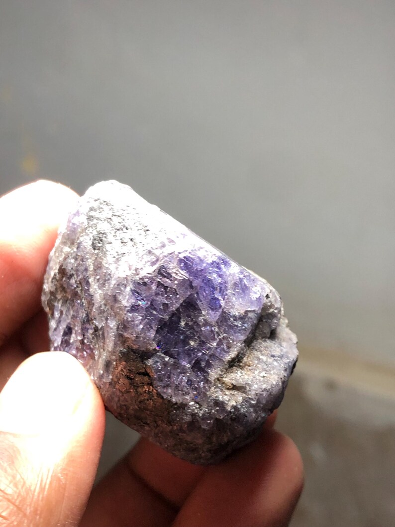 May include: A rough-cut, natural gemstone with a mix of gray and purple hues. The stone has a crystalline structure with visible facets and a textured surface. The color ranges from deep violet to lighter shades, with areas of gray.