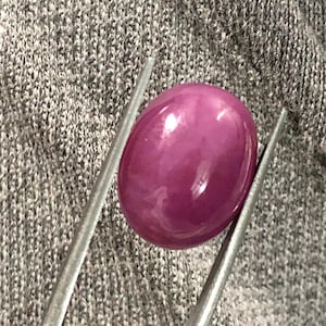 Puede incluir: Una gema pulida, de forma ovalada y de color rojo púrpura, es sostenida por unas pinzas metálicas sobre un fondo de tela gris. La gema tiene una superficie lisa y reflectante, y las pinzas están posicionadas para sujetarla de forma segura.
