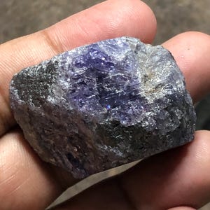 May include: A rough, uncut gemstone with a mix of dark gray and vibrant purple hues. The stone has a crystalline structure with visible facets and a slightly reflective surface. The overall shape is irregular, showcasing its natural, raw form.