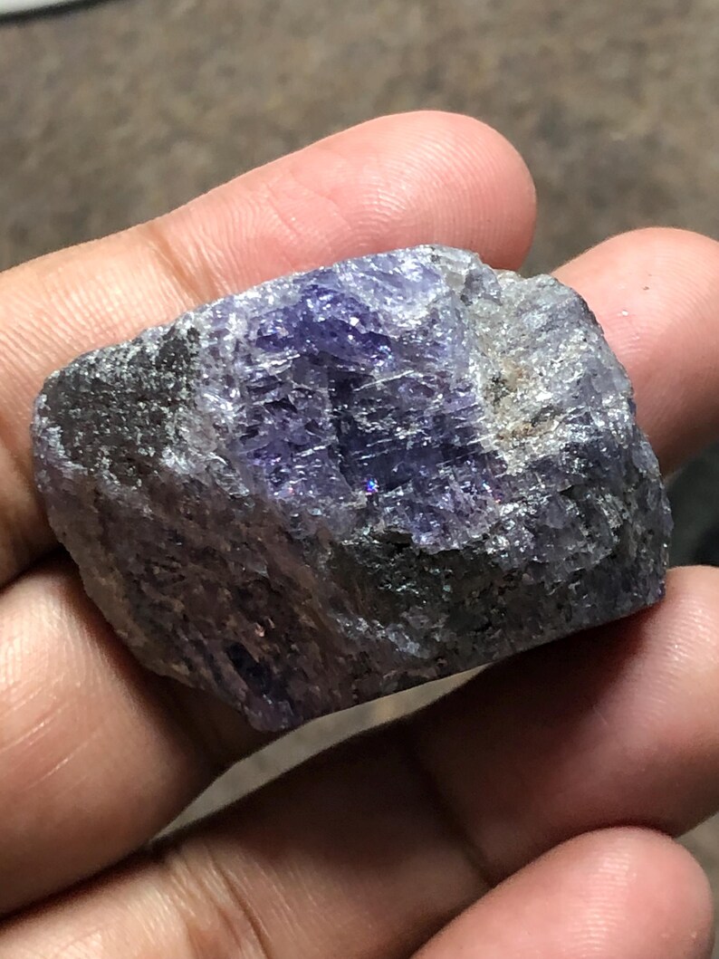 May include: A rough, uncut gemstone with a mix of dark gray and vibrant purple hues. The stone has a crystalline structure with visible facets and a slightly reflective surface. The overall shape is irregular, showcasing its natural, raw form.