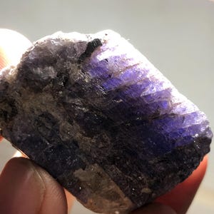May include: A rough-hewn, natural stone specimen with a striking display of color. The stone features a blend of deep purple and gray hues, with areas of intense violet. The texture appears coarse and crystalline, with visible mineral formations.