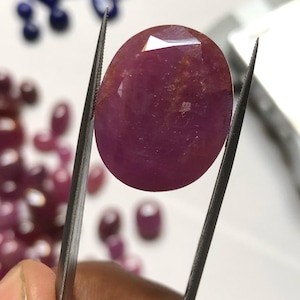 May include: A vibrant pink oval-shaped gemstone with a faceted cut. The gemstone is held by tweezers and appears to be a ruby.