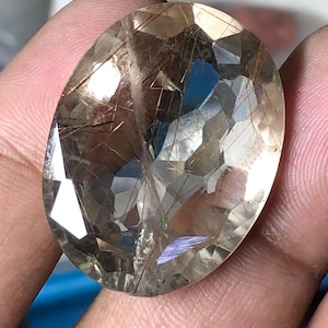 Pode incluir: Uma pedra preciosa oval com uma superfície facetada. A gema é de cor âmbar claro com inclusões internas castanho-douradas. As facetas criam um jogo de luz e sombra, realçando o brilho da gema. A pedra preciosa é segurada em uma mão.
