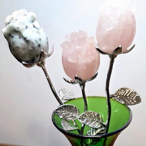 May include: A decorative arrangement of three carved stone roses in a green glass vase. The roses are made of different stones, including a speckled gray and white stone and two pink rose quartz stones. The stems and leaves are silver-colored metal.