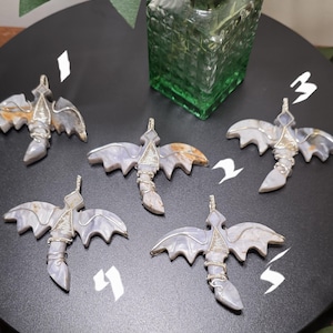 May include: Five gemstone bat pendants with silver wire wrapping, displayed on a black surface. The bats have gray and tan wings and bodies. A green glass vase with greenery is in the background.