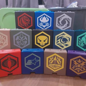 May include: A collection of colorful, square boxes with hexagonal designs. Each box features a unique emblem in various colors, including red, blue, and yellow. The boxes are arranged in rows, showcasing a variety of graphic symbols and patterns.