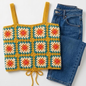 May include: A mustard yellow crochet top with a square pattern in orange, teal, and cream colours. The top has shoulder straps and a tie at the waist. Paired with blue denim jeans.
