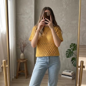 May include: Mustard yellow crochet top with short sleeves and a textured openwork design. The top is paired with light blue jeans. The image is taken in front of a mirror.
