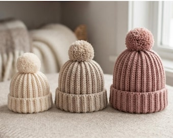 May include: Three crochet baby beanies in cream, beige, and dusty rose, each featuring a ribbed texture and a matching pom-pom on top. The text CROCHET BABY BEANIE PATTERN PREEMIE → CHILD is displayed above the hats in black capital letters.