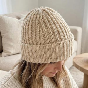 May include: A cream-colored knit beanie featuring a thick, vertical ribbed texture and a wide, folded cuff. The hat is constructed from a chunky yarn, creating a consistent, repeating pattern that extends from the crown down to the brim.