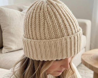Peut inclure: Un bonnet en tricot de couleur crème présentant une texture côtelée verticale épaisse et un large revers replié. Le bonnet est confectionné avec une laine épaisse, créant un motif répétitif cohérent du sommet jusqu'au bord.