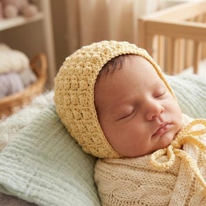 May include: A pale yellow crochet baby bonnet with a textured bobble stitch pattern and matching yellow yarn ties, worn by a sleeping infant wrapped in a cream-colored cable knit blanket, resting on a soft light green textured fabric.