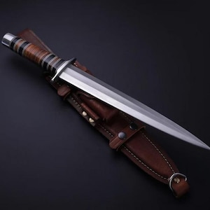 May include: A fixed-blade knife with a double-edged steel blade and a stacked leather handle featuring black and silver accents. The knife is positioned diagonally across a brown leather sheath that has visible stitching and metal snap fasteners.