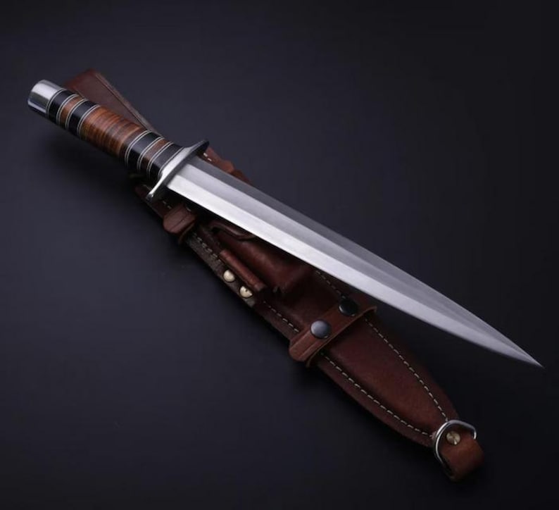 May include: A fixed-blade knife with a double-edged steel blade and a stacked leather handle featuring black and silver accents. The knife is positioned diagonally across a brown leather sheath that has visible stitching and metal snap fasteners.