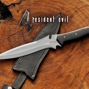 May include: A silver-bladed knife with a black handle and gold rivets, on a black leather sheath. The knife is labelled "Resident Evil" and is set against a wooden background.