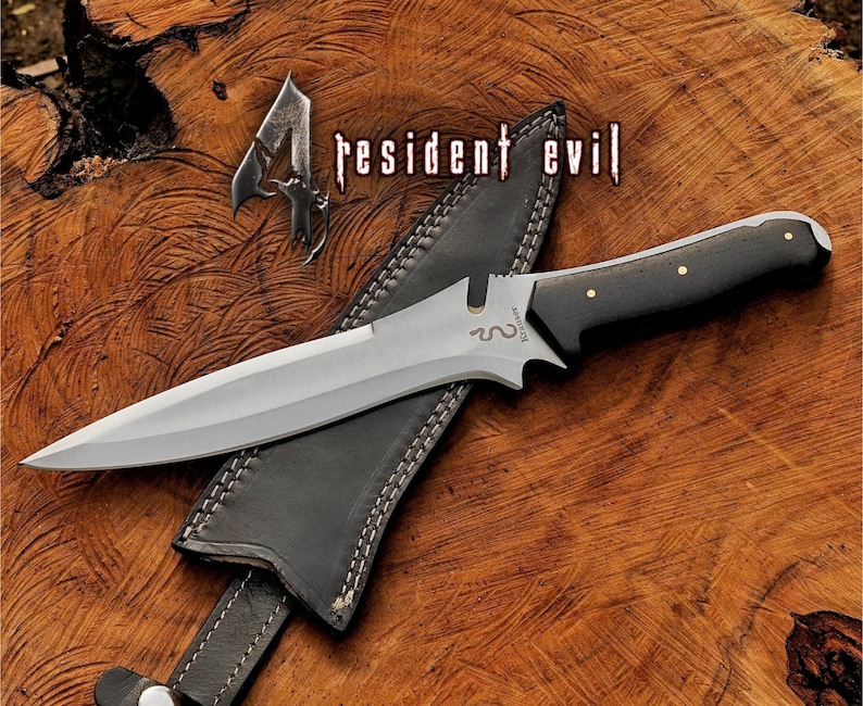 May include: A silver-bladed knife with a black handle and gold rivets, on a black leather sheath. The knife is labelled "Resident Evil" and is set against a wooden background.