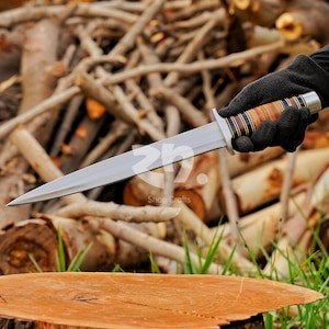 May include: A double-edged fixed blade knife with a pointed steel tip and a central ridge. The handle features alternating bands of light brown wood, dark brown wood, and black material, finished with a silver-colored metal pommel and guard.