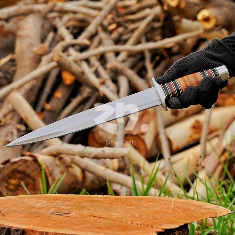 May include: A double-edged fixed blade knife with a pointed steel tip and a central ridge. The handle features alternating bands of light brown wood, dark brown wood, and black material, finished with a silver-colored metal pommel and guard.