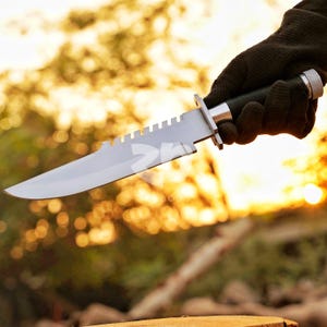 May include: A large, silver survival knife with a serrated edge and a green handle. The knife is held in a gloved hand against a blurred background of trees and sunlight. The blade is approximately 12 inches long.