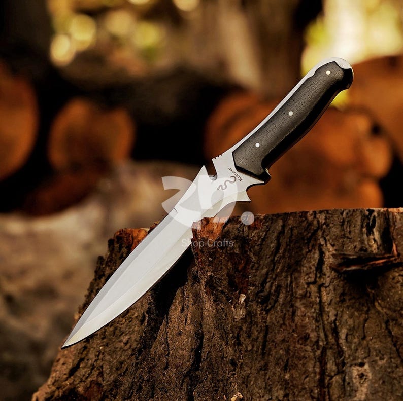 May include: A stainless steel knife with a black handle, resting on a wooden surface. The blade is sharp and pointed, with a decorative design near the handle. The handle has three silver rivets. The knife is a hunting or survival tool.