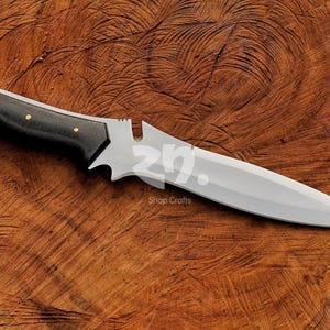 May include: A handcrafted knife featuring a polished silver blade and a black handle with small gold rivets. The knife rests on a textured wooden surface. The text "Shop Crafts" is visible. A decorative item.