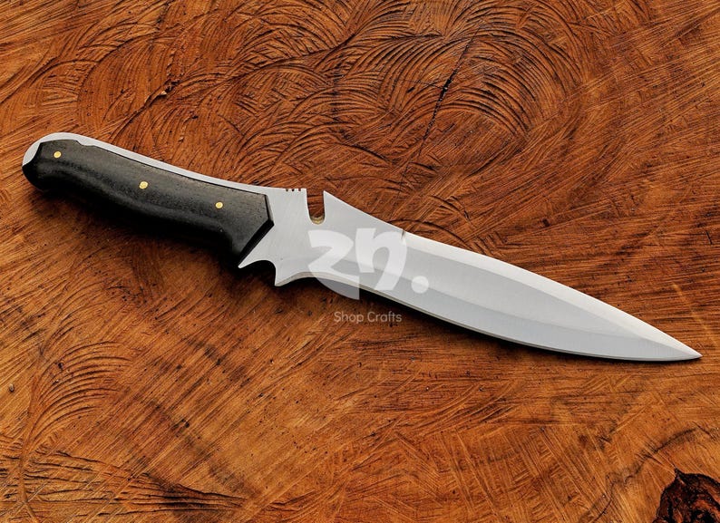 May include: A handcrafted knife featuring a polished silver blade and a black handle with small gold rivets. The knife rests on a textured wooden surface. The text "Shop Crafts" is visible. A decorative item.