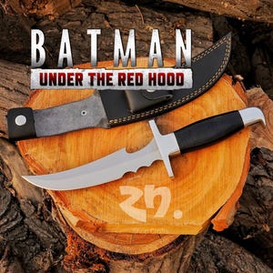 May include: A stainless steel knife with a black handle and a leather sheath. The knife has a serrated edge and a crossguard. The text "BATMAN UNDER THE RED HOOD" is displayed above the knife.
