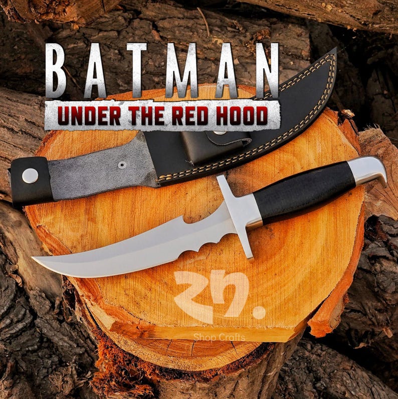 May include: A stainless steel knife with a black handle and a leather sheath. The knife has a serrated edge and a crossguard. The text "BATMAN UNDER THE RED HOOD" is displayed above the knife.