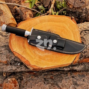 May include: A black hunting knife with a silver bolster and a black handle, sheathed in a black leather case with white stitching. The knife rests on a wooden surface. The text "Shop Crafts" is visible.