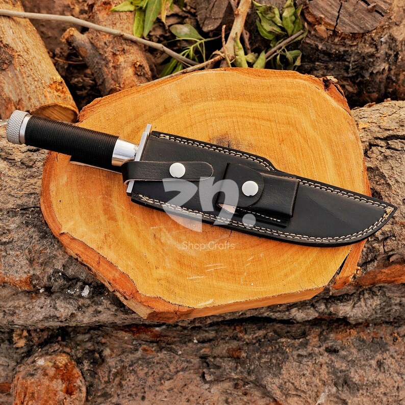 May include: A black hunting knife with a silver bolster and a black handle, sheathed in a black leather case with white stitching. The knife rests on a wooden surface. The text "Shop Crafts" is visible.