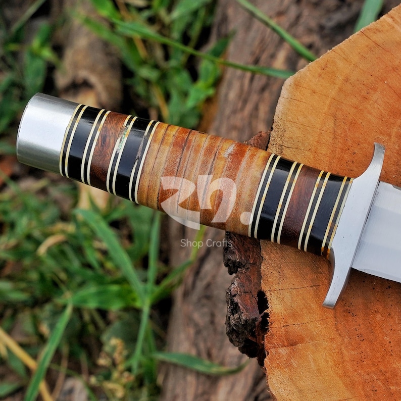 May include: A knife handle featuring alternating segments of polished dark wood and light tan wood, separated by thin black and white spacers. The handle includes a silver-colored metal pommel at the end and a silver-colored metal guard near the blade.